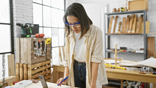 A young woman wearing safety glasses marks wood in a well-equipped carpentry workshop.