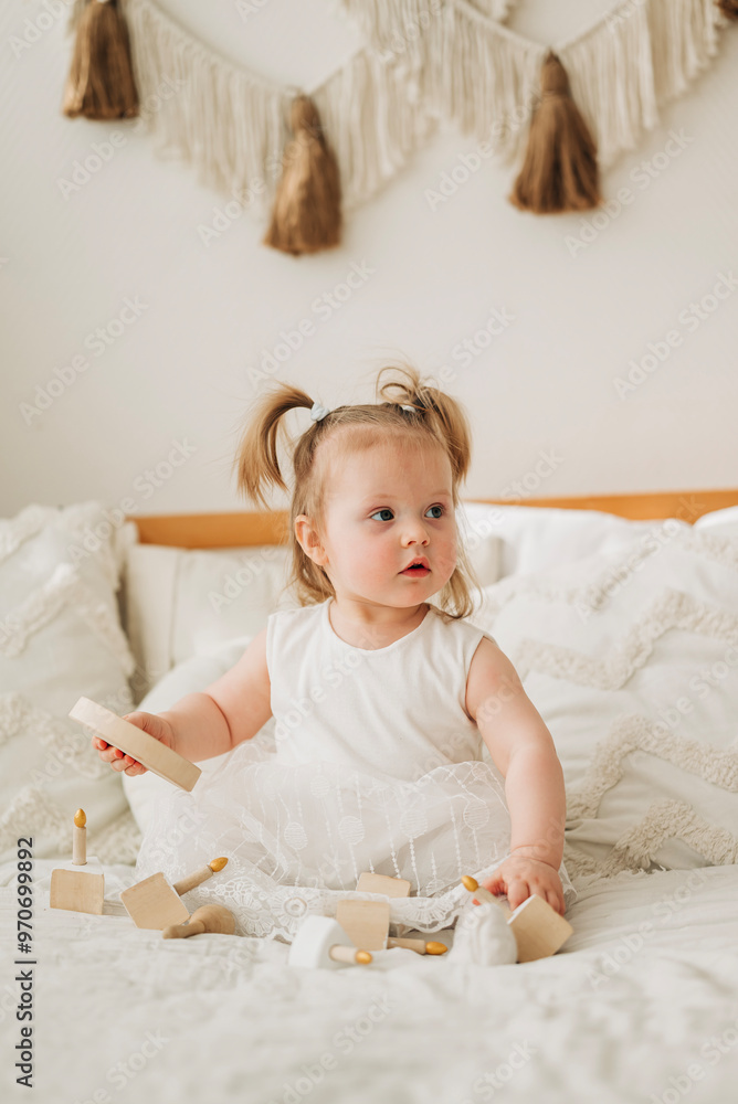 Fototapeta premium Cute beautiful girl baby sitting on the bed in the bedroom and playing in eco wooden toys cubes and pyramid