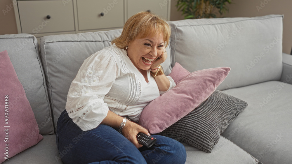 Middle-aged blonde woman laughing and holding a tv remote while sitting on a comfortable sofa in a cozy living room. Middle-aged blonde woman laughing and holding a tv remote while sitting on a comfortable sofa in a cozy living room.