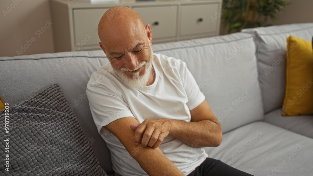 Middle-aged man with grey hair scratching his arm, sitting comfortably in a modern living room with a cozy sofa and colorful cushions, suggesting a relaxed home environment. Middle-aged man with grey hair scratching his arm, sitting comfortably in a modern living room with a cozy sofa and colorful cushions, suggesting a relaxed home environment.