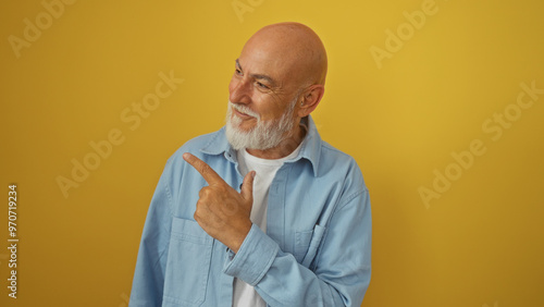 Fotografie Handsome mature bald man with a grey beard and blue shirt standing isolated against a bright yellow wall while smiling and pointing to the side
