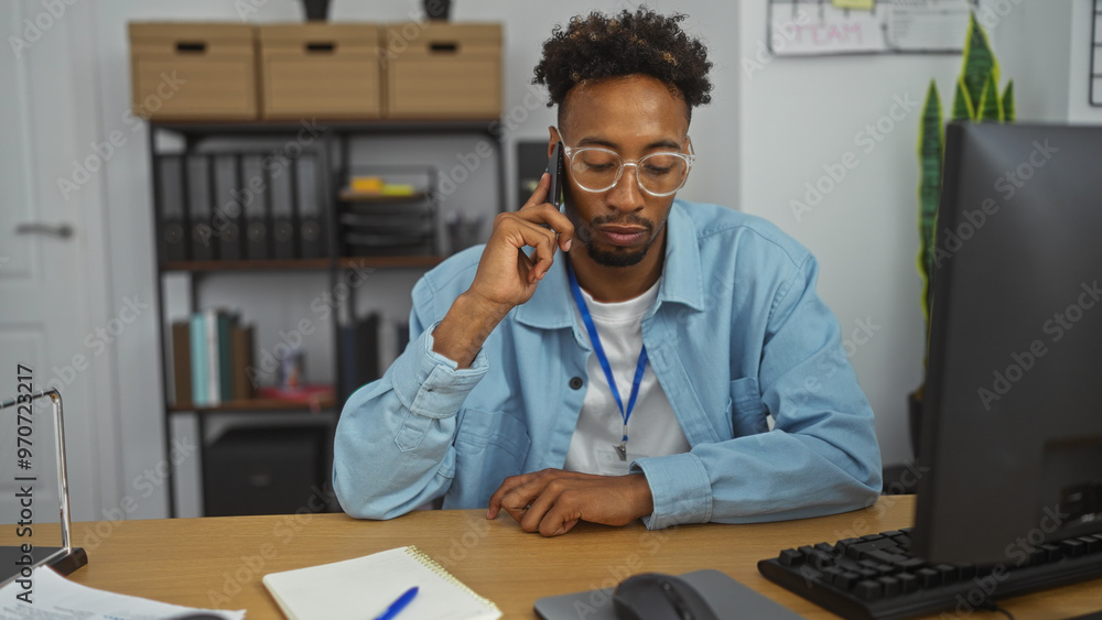 Young handsome african american man with a beard working in an office, seated at a desk on a phone call, surrounded by office supplies and a computer in an indoor workplace setting. Young handsome african american man with a beard working in an office, seated at a desk on a phone call, surrounded by office supplies and a computer in an indoor workplace setting.