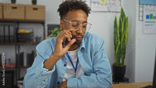 Handsome young african american man with a beard and glasses working in an office wearing a blue shirt, surrounded by an indoor office setting with plants and shelves.