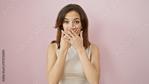 Surprised young hispanic woman covering mouth with hands against a pink isolated background.