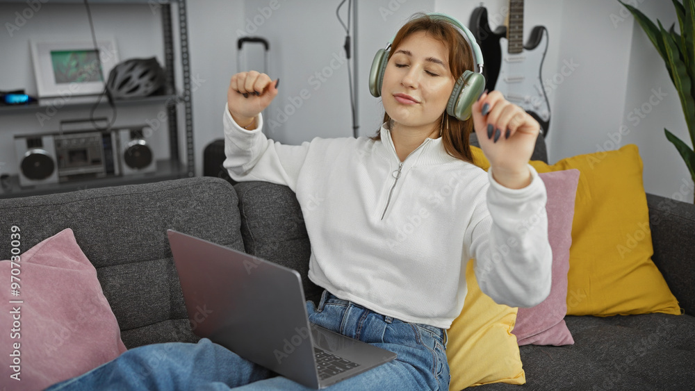 Obraz premium A joyful young woman wearing headphones dances to music while using a laptop in a cozy living room.