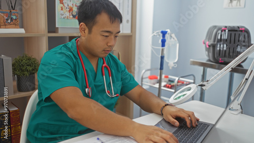 A young asian male veterinarian, wearing green scrubs and a red stethoscope, is focused on his laptop in a modern veterinary clinic.