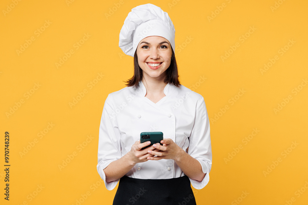 Young chef cook baker woman wears white shirt black apron uniform toque chefs hat hold in hand use mobile cell phone look for recipe isolated on plain yellow background studio. Cooking food concept. Young chef cook baker woman wears white shirt black apron uniform toque chefs hat hold in hand use mobile cell phone look for recipe isolated on plain yellow background studio. Cooking food concept.