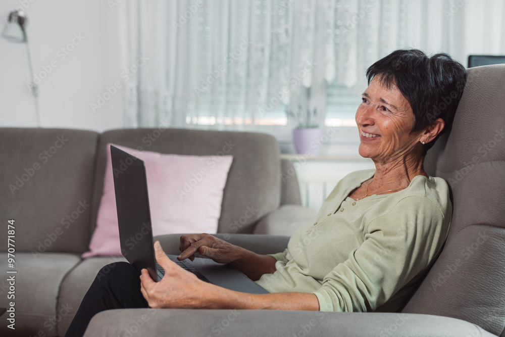 A senior woman sitting comfortably in a chair, smiling while using a laptop. The room is well-lit with a cozy atmosphere, featuring a fitness bike in the background. A senior woman sitting comfortably in a chair, smiling while using a laptop. The room is well-lit with a cozy atmosphere, featuring a fitness bike in the background.