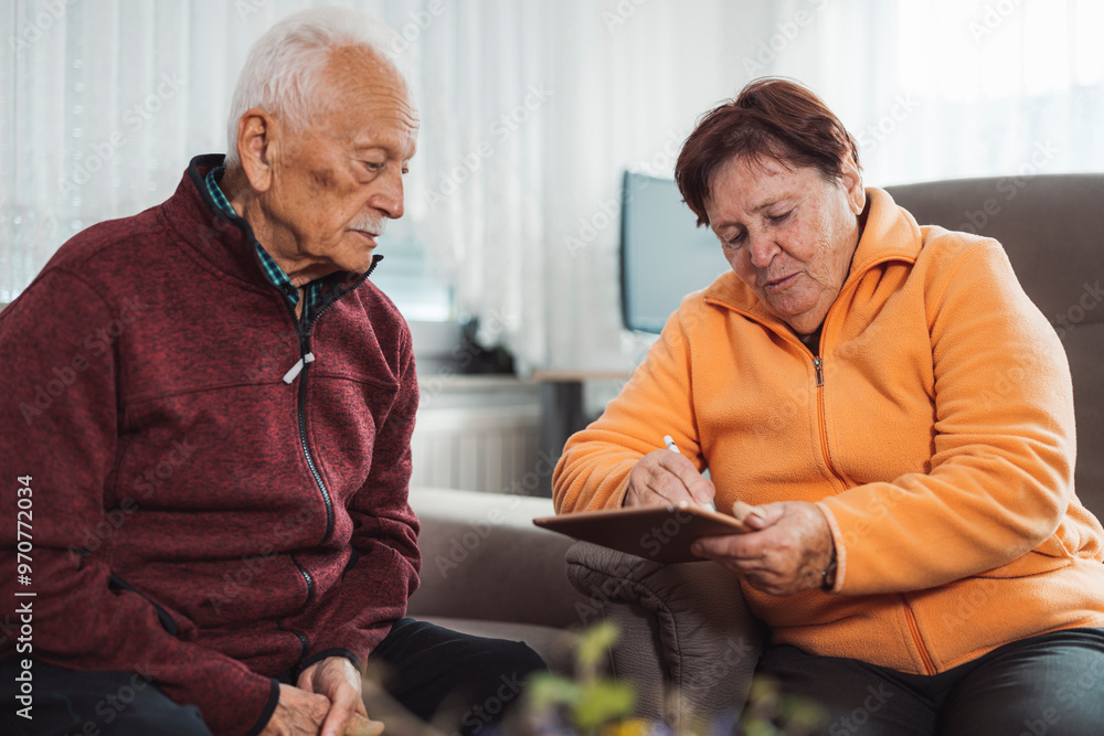 An elderly couple sitting on a couch, engaged in a conversation while looking at a tablet. The man has gray hair and a mustache, wearing a red sweater. The woman has short brown hair, wearing an orang An elderly couple sitting on a couch, engaged in a conversation while looking at a tablet. The man has gray hair and a mustache, wearing a red sweater. The woman has short brown hair, wearing an orang