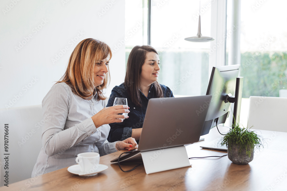 Two women working together at a modern office desk, one holding a glass of water and smiling, while the other is focused on a laptop. A cup and a small plant are on the table. Two women working together at a modern office desk, one holding a glass of water and smiling, while the other is focused on a laptop. A cup and a small plant are on the table.