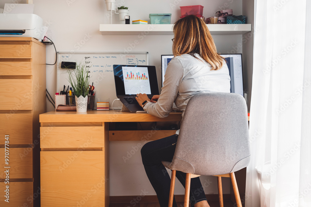 A woman working at a desk with two computer screens displaying data and graphs. She is focused on her work, holding glasses in her hand. The workspace is modern and organized, with a plant and whitebo A woman working at a desk with two computer screens displaying data and graphs. She is focused on her work, holding glasses in her hand. The workspace is modern and organized, with a plant and whitebo