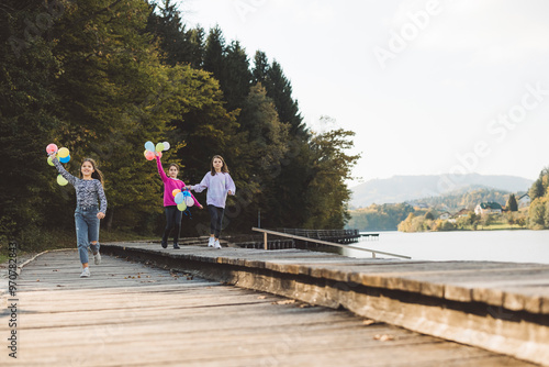 Three girls joyfully running along a wooden pier by a lake, holding colorful balloons. The scene is set in a serene natural environment with trees and hills in the background, under a clear sky.