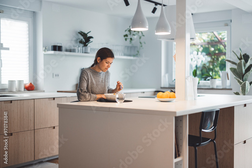 A young woman sitting at a dining table, enjoying a bowl of salad. She has long brown hair tied back and is focused on her meal. The setting is modern with a minimalist design, featuring a vase of flo