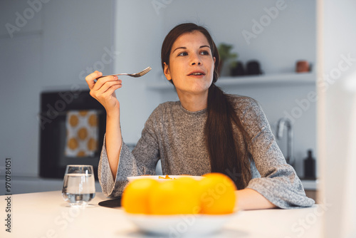 A young woman sitting at a dining table, enjoying a bowl of salad. She has long brown hair tied back and is focused on her meal. The setting is modern with a minimalist design, featuring a vase of flo