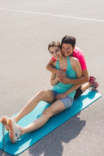 A young woman sitting on an exercise mat, holding a water bottle, while an older woman embraces her from behind, both smiling. They are outdoors on a sunny day, suggesting a fitness or training sessio photo