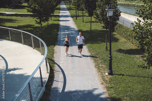 A couple jogging together on a scenic path surrounded by greenery. The path is lined with trees and a lamp post, creating a peaceful atmosphere. The sun casts shadows on the ground, enhancing the outd photo