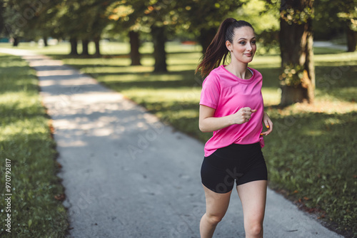 A young woman jogging in a park, wearing a pink t-shirt and black shorts. She has a ponytail and is smiling, surrounded by trees and a path.