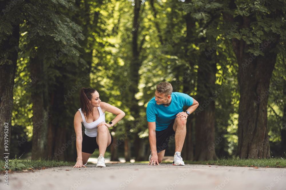 A man and woman in athletic attire are positioned in a starting stance on a path surrounded by trees. The woman is on her knees, while the man is in a crouched position, both ready to race. The scene