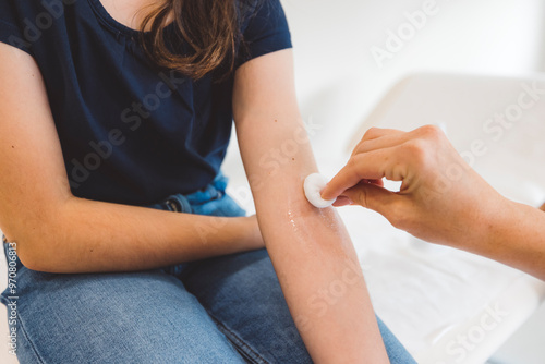 A young girl sitting on a medical examination table, looking concerned as a healthcare professional examines her arm. Another woman stands nearby, observing the situation. The setting is bright and cl