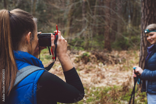 A woman taking a photo of another woman in a forest setting. The photographer is wearing a blue vest and has long hair tied back. The subject is smiling and wearing a headlamp, standing with trekking photo