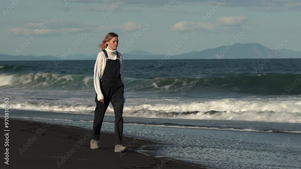Full length view of female fashion model struts along black sandy beach against background of ocean waves during summer vacation. Woman rocking sweater, denim jumpsuit and trainers. Handheld real time Full length view of female fashion model struts along black sandy beach against background of ocean waves during summer vacation. Woman rocking sweater, denim jumpsuit and trainers. Handheld real time