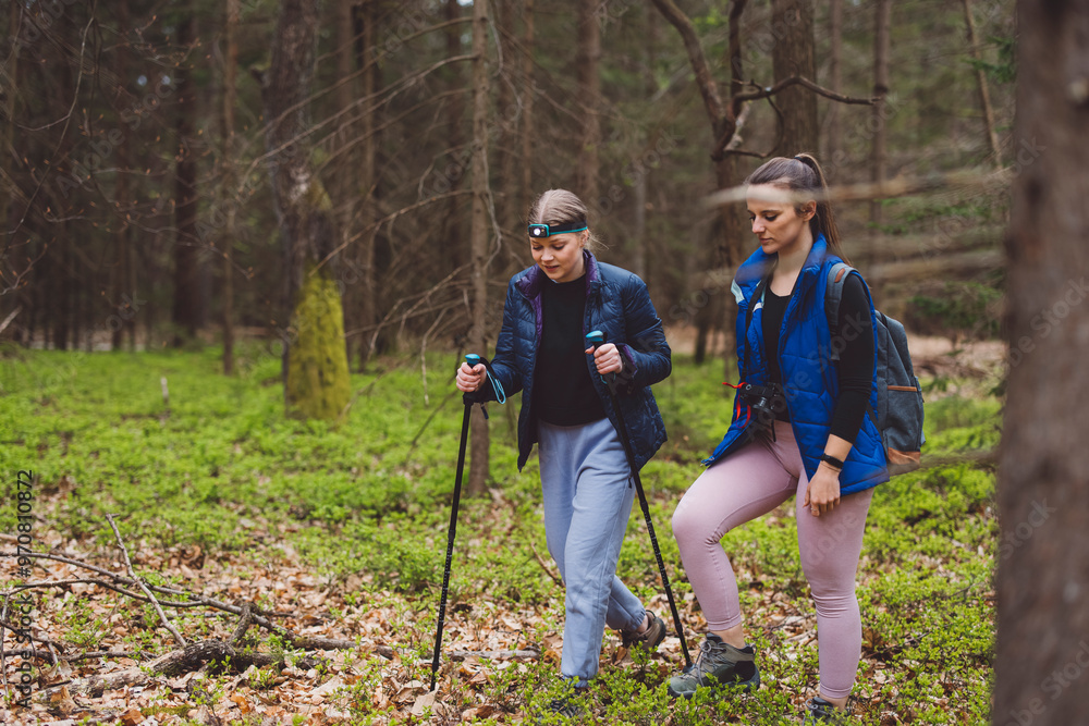 Two women hiking in a forest, one using trekking poles and wearing a ...
