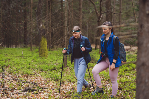 Two women hiking in a forest, one using trekking poles and wearing a headlamp, the other carrying a backpack. They are surrounded by green foliage and trees.