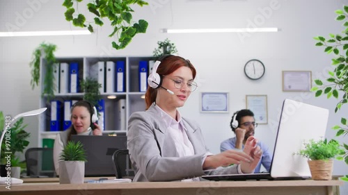 international company, young woman with headset and glasses works in call center and answers calls sitting at computer against background of colleagues in office photo