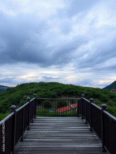 A wooden deck with a black railing overlooking a lush green hillside under a cloudy sky.