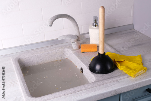 A plunger and yellow rubber gloves on top of a clogged kitchen sink