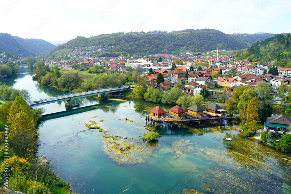Fototapeta premium View from the fortress in the Old Town to the centre of Bosanska krupa: clear waters of the Una River, fishermen's houses on stilts, low-rise buildings and forested mountains in the background.