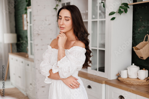 Women's day. Romantic attractive young woman 20-25 years with long curly hair in white dress is poses with crossed hands and looks at camera at modern stylish interior of home kitchen on background.