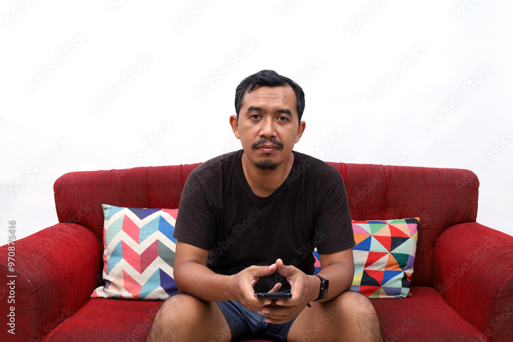 Obraz premium Funny middle age asian man sitting on the red sofa looking at the camera, doing nothing. Isolated on white