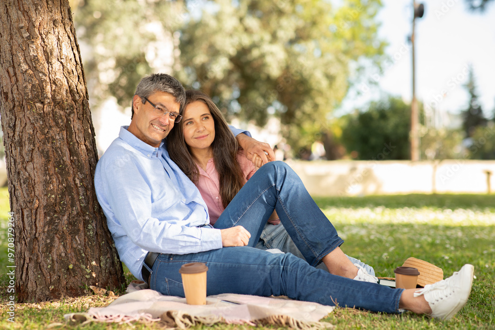 A couple sits together on a blanket, leaning against a tree in a lush park. They share a warm moment, smiling while sipping coffee, enjoying the sunny day. A couple sits together on a blanket, leaning against a tree in a lush park. They share a warm moment, smiling while sipping coffee, enjoying the sunny day.