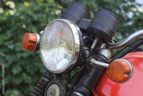 part of an old motorcycle with a white glass headlight and black handlebars near a red gas tank on a summer street