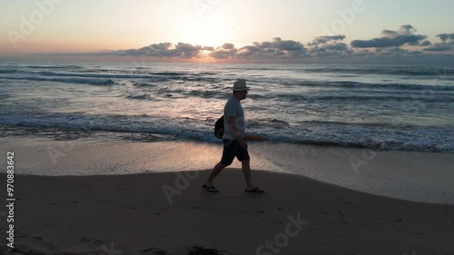 Man strolls on the beach at sunrise, carrying an orange beach umbrella and black bag. Wearing a white hat, light shirt, shorts, and flip-flops. Slow motion. photo