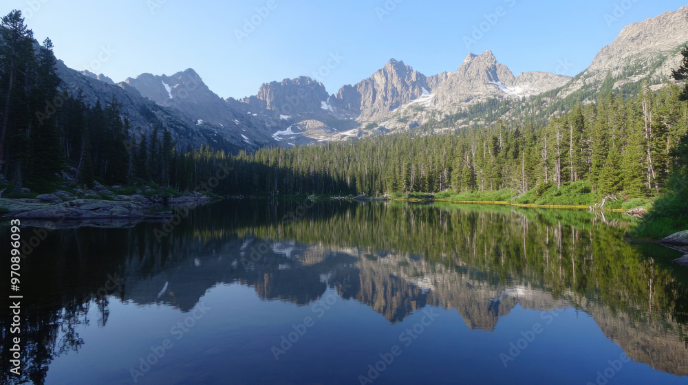 Fototapeta premium A serene alpine lake reflecting towering mountains and dense pine forests in the early morning light.