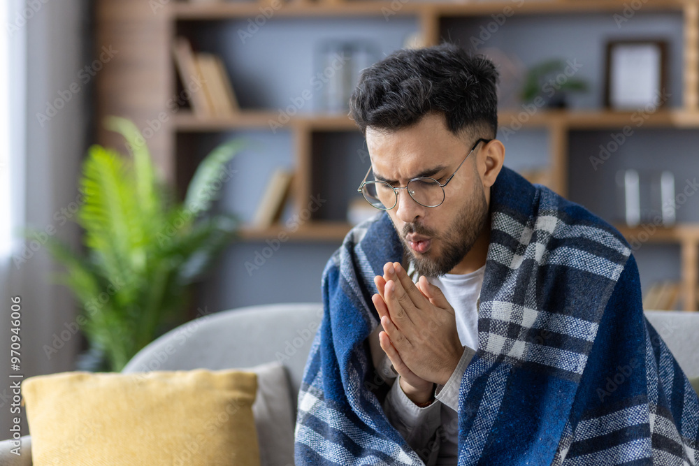 Naklejka premium A young Indian man sits at home on the sofa covered with a blanket, rubbing and blowing air on his hands to keep warm. Suffering from flu and cold