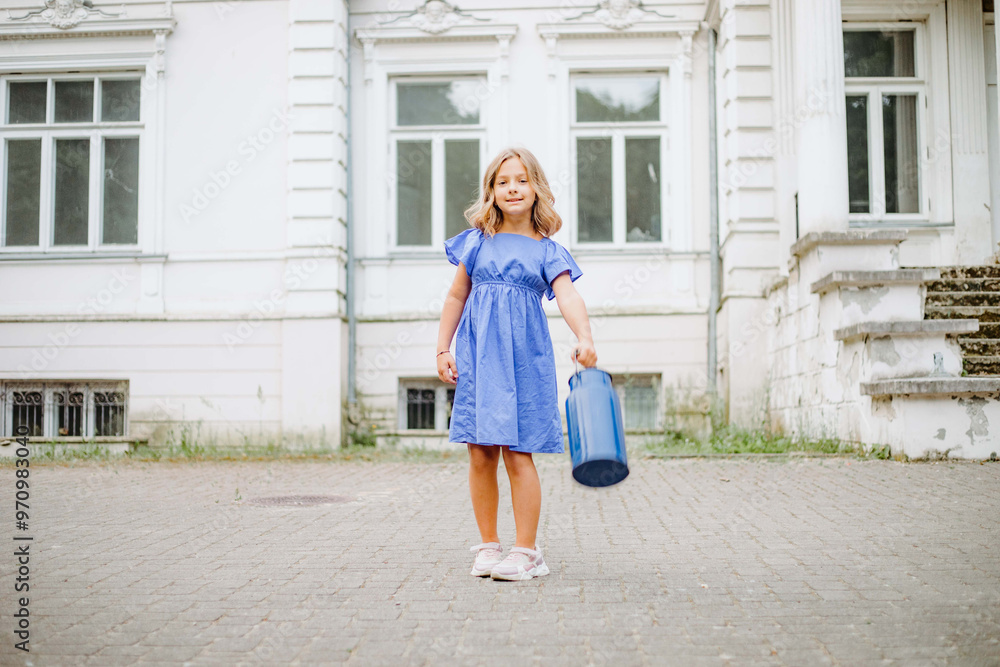 Naklejka premium A little girl in a blue dress holding a blue bucket near a white building
