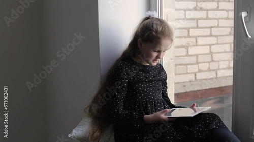 A portrait of a young girl sits on the windowsill and enthusiastically reads on a tablet. A Caucasian girl sits on the windowsill with a tablet and uses it. Teenager learning lessons at home