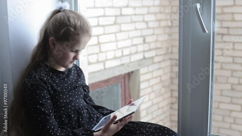 A young girl sits on the windowsill and enthusiastically reads on a tablet. Distance learning concept. Teenager learns lessons at home. A girl in a dress sits on the windowsill with a tablet 