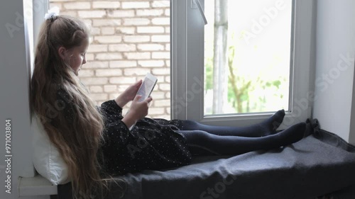 A Caucasian girl with light hair sits by the window and reads on a tablet. Development and learning of children remotely
