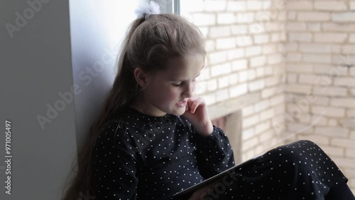 A portrait of a young girl sits on the windowsill and enthusiastically reads on a tablet. A Caucasian girl sits on the windowsill with a tablet and uses it. Teenager learning lessons at home