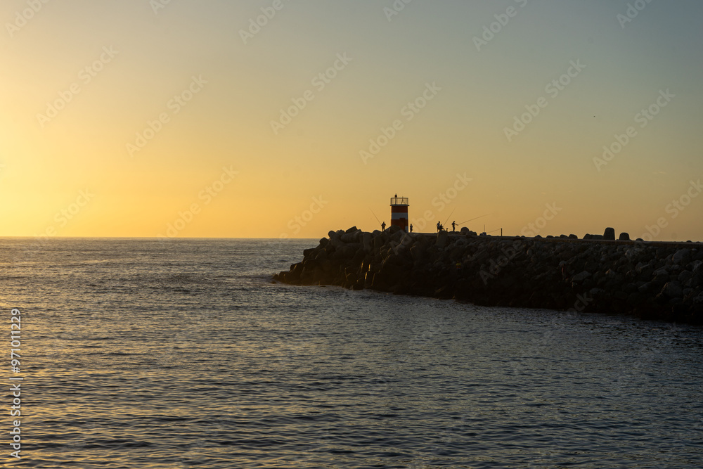 Fototapeta premium A breathtaking sunset at Nazaré, Portugal, with the sun dipping below the horizon, framed perfectly between two lighthouses at the entrance of the marina, casting golden reflections on the sea.
