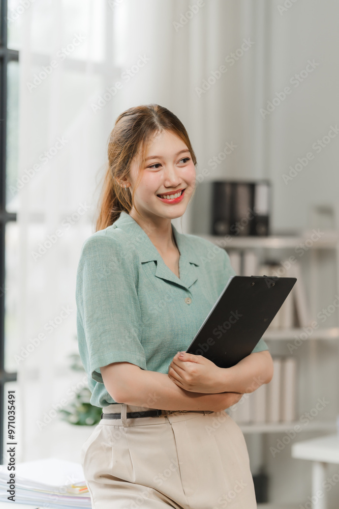 Confident Businesswoman: A young, professional woman in a mint green blouse and beige pants stands confidently in her office, holding a black clipboard, radiating positivity and competence.  