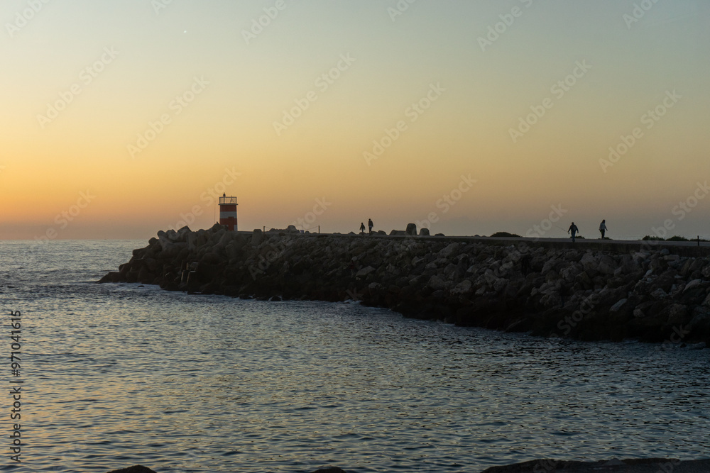 Silhouettes of people fishing on the pier by the Nazaré lighthouse at sunset, the vibrant orange sky reflecting over the ocean, capturing a peaceful coastal moment. Silhouettes of people fishing on the pier by the Nazaré lighthouse at sunset, the vibrant orange sky reflecting over the ocean, capturing a peaceful coastal moment.