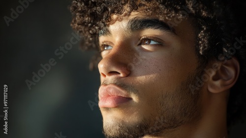 A close-up portrait of a young man with curly hair, looking away thoughtfully, natural light hitting his face