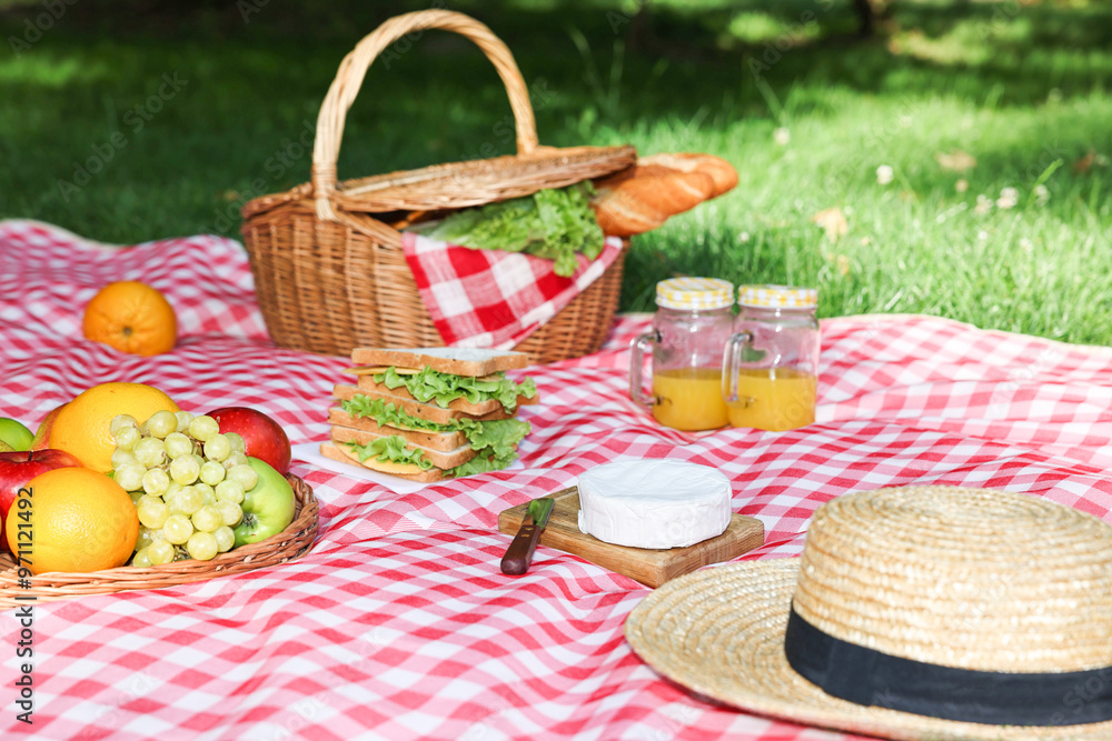 Picnic wicker basket with delicious food, drink and tableware on checkered blanket outdoors Picnic wicker basket with delicious food, drink and tableware on checkered blanket outdoors