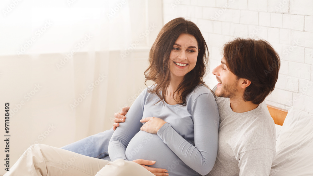 Happy pregnant couple relaxing on bed at home together, cuddling and talking, free space Happy pregnant couple relaxing on bed at home together, cuddling and talking, free space