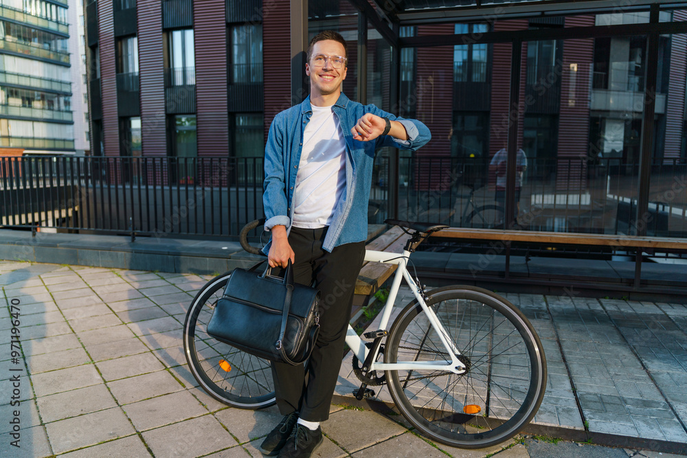 Obraz premium A smiling man stands with his bicycle, checking his watch, dressed casually, holding a briefcase.